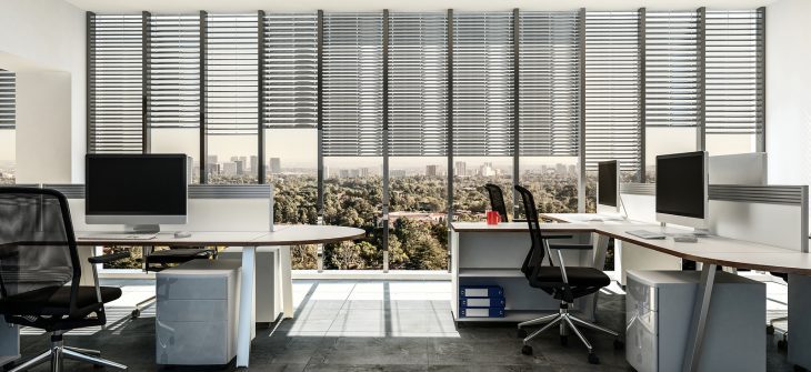 Modern business office with multiple workstations around table style desks with cabinets, tiled grey floor and large windows with blinds. 3d Rendering.